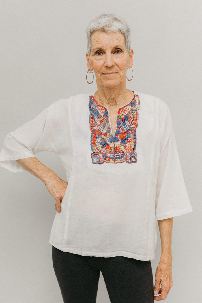 Older woman in short version of Egyptian top. Top is made in a white gauze fabric with multi-colored embroidery on yoke. Model has one hand on hip and poses with white backdrop.