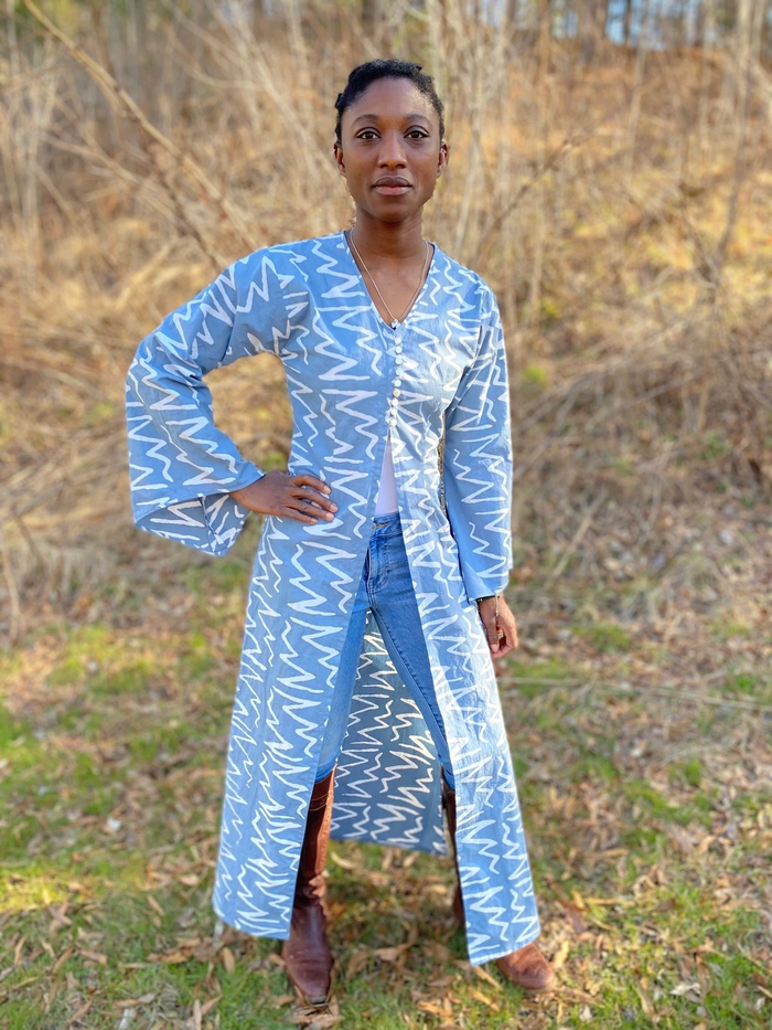 Black woman wearing a long blue/grey batiked entari with brown boots. She is standing outside.