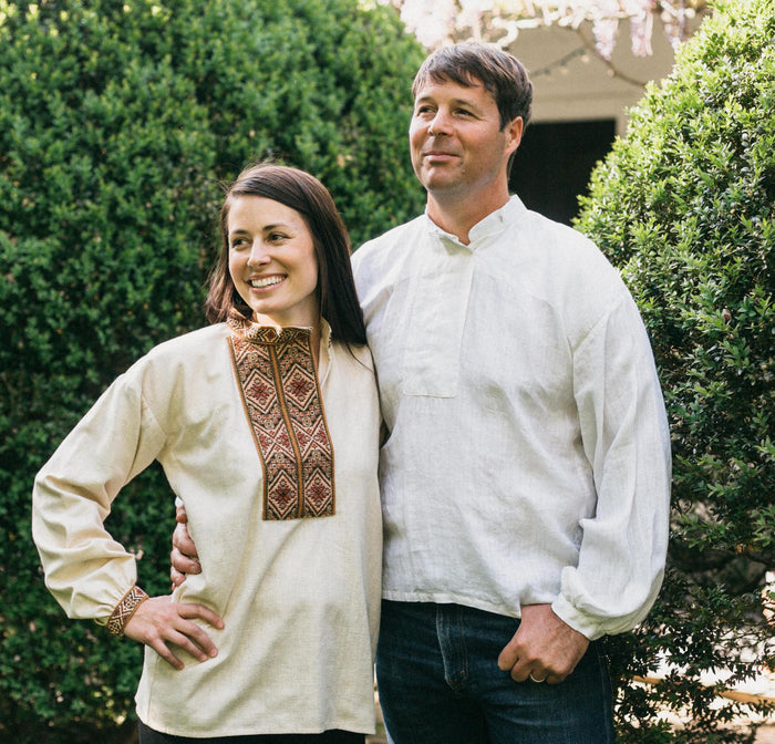 Woman and man standing outside with hedges behind them. Woman is wearing embroiders View B and man is wearing plain white View B. The are standing side by side with arms around eachother.