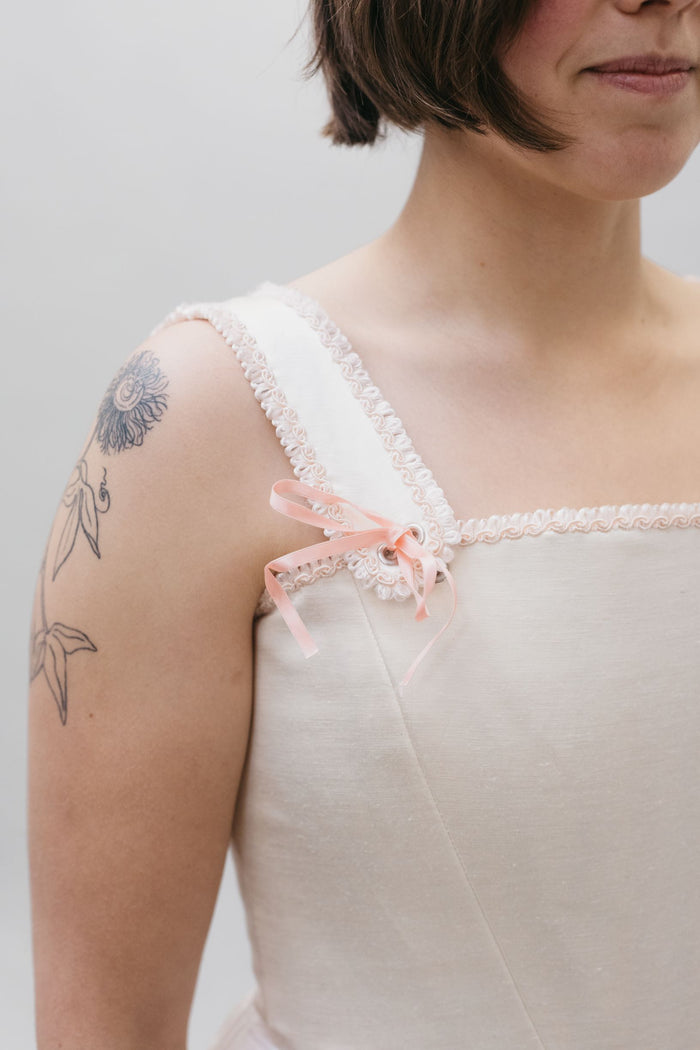 Close up of the square neck Lady's Corset strap withe a pink bow ribbon at the base of the strap on white woman with dark hair and tattoo. Ribbon tie detail for Corset top.