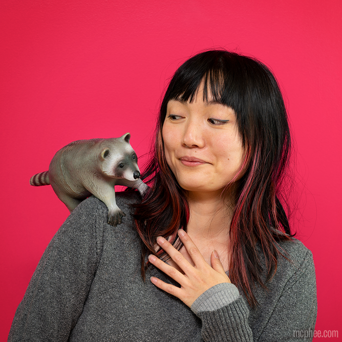 A woman smiles at a Baby Raccoon Buddy with posable paws playfully adjusted on her shoulder, set against a vibrant pink background.