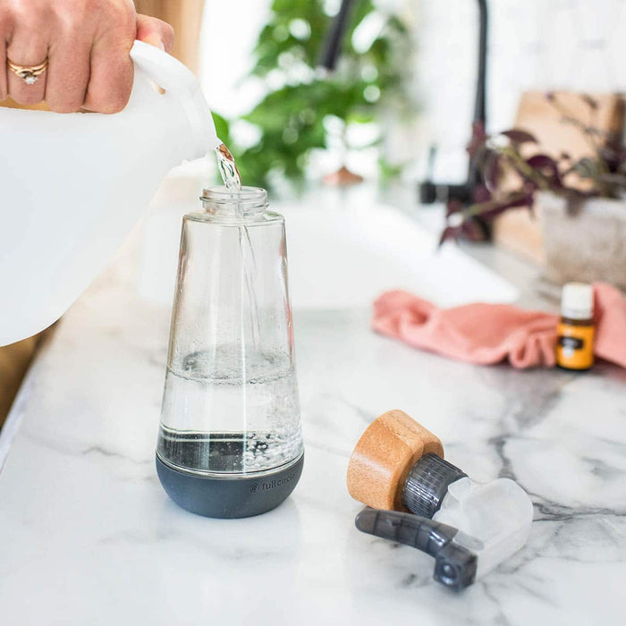 Glass bottle with lid off while a person pours in water, styled on a marble counter with cleaning cloth & essential oils in the background.