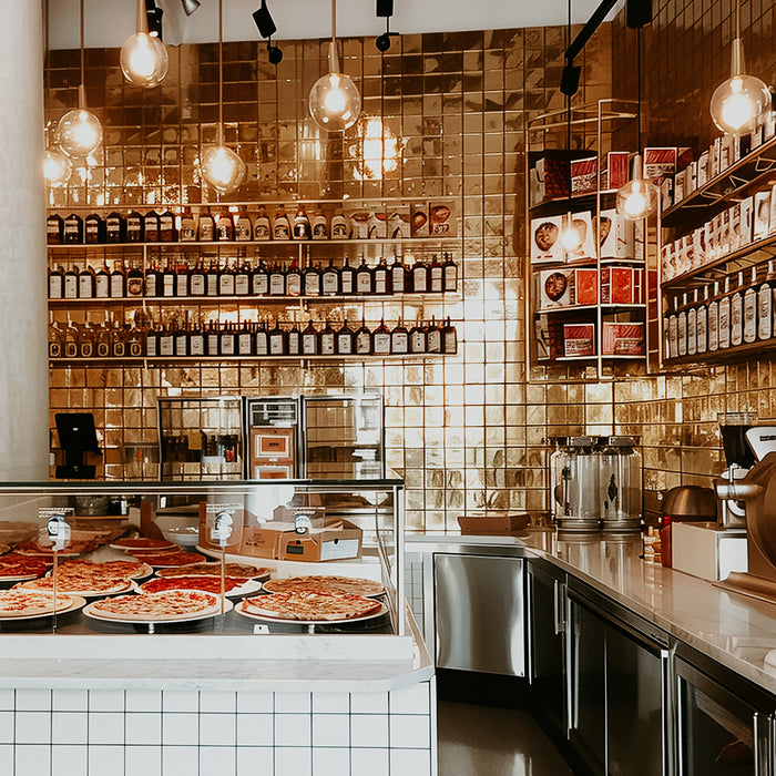 Interior view of a pizzeria with pizzas displayed in the foreground and shelves of ingredients and equipment in the background.