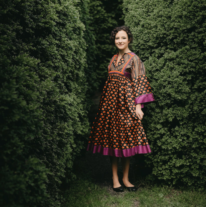 Young woman standing between two bushed with floral poppy print Afghan Nomad dress.