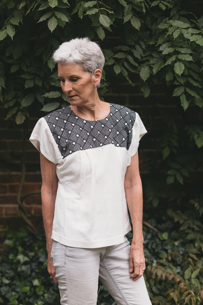 Older woman wearing Old Mexico Blouse outdoors in front of a brick wall with wisteria vines growing behind her.