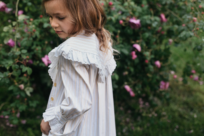 Back side view of little brunette girl looking down wearing Childhood Dreams close up of wide triangular ruffled neckline. Surrounded by greenery and purple roses.