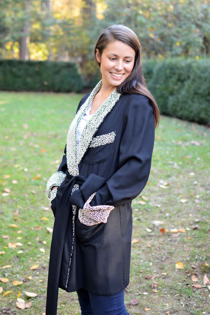 Young brunette white woman smiling down turned to the left and standing surrounded by greenery wearing the 238 Le Smoking Jacket with hands in pockets with quilted lapel and cuffs and pocket band, with sash wrapped around the waist.