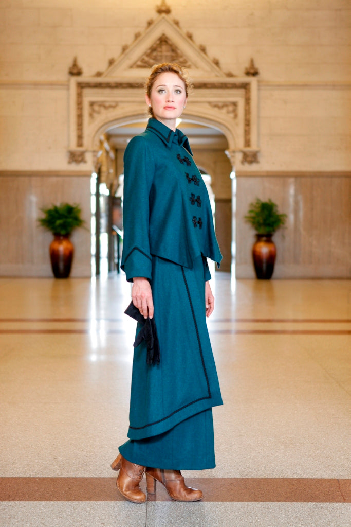 Young blonde woman standing in a lobby wearing the Metropolitan Suit holding black gloves in her right hand and brown heeled boots on her feet.