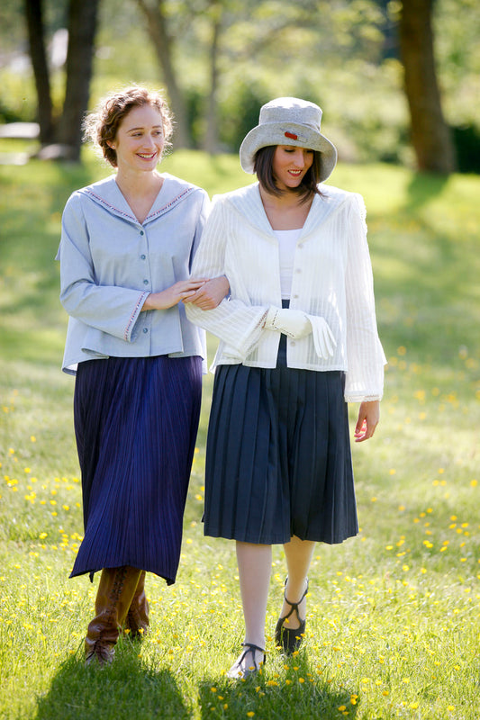 Two  women walking linking arms, surrounded by greenery. Woman on the left wearing #270 Metro Middy Blouse in blue and long skirt. Woman on the right is wearing a hat with the Metro Middy Blouse in white and a knee length skirt.
