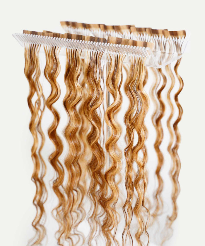 Several rows of wavy, light brown hair extensions elegantly displayed on a transparent rack against a white background.