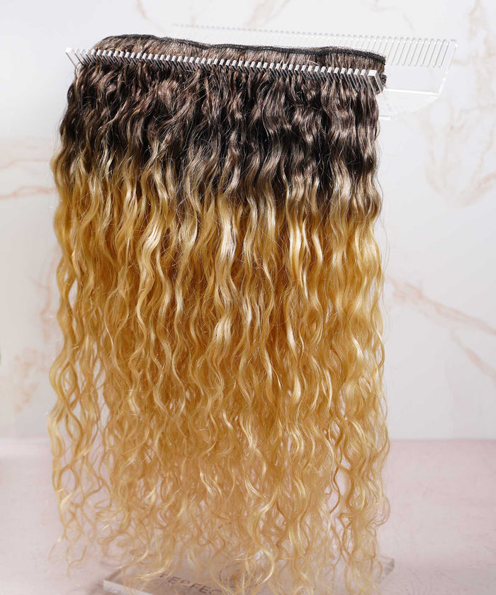 A bundle of long, wavy hair extensions with dark brown roots fading to light blonde ends, displayed on a clear stand, light background.