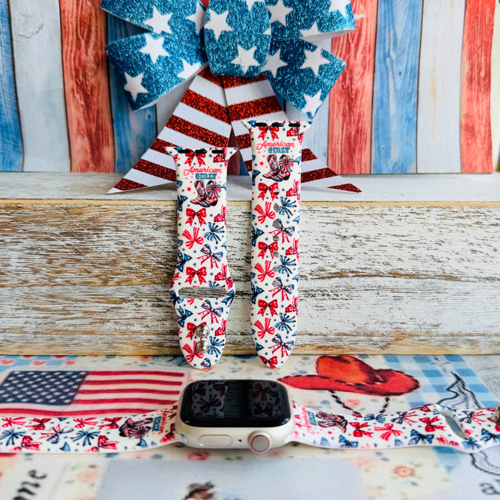 American Girly silicone Apple Watch band with red and blue coquette bows, mini cowboy boots, stars, and "American Girly" script on a white base, shown displayed and worn on Apple Watch against a patriotic 4th of July backdrop.