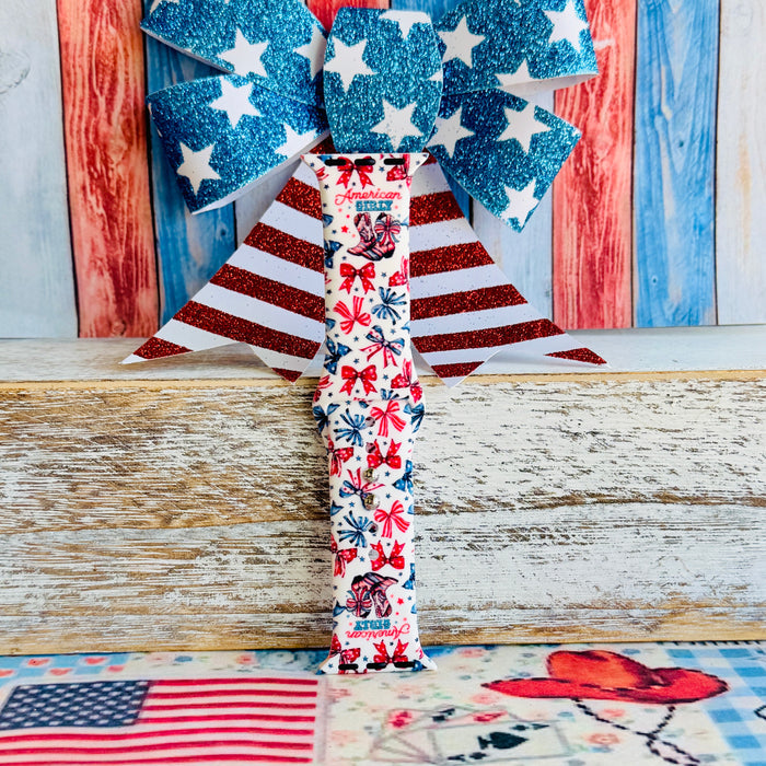 American Girly silicone Apple Watch band with red and blue coquette bows, mini cowboy boots, stars, and "American Girly" script on a white base, shown displayed and worn on Apple Watch against a patriotic 4th of July backdrop.
