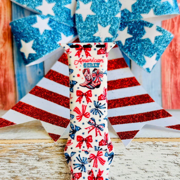 American Girly silicone Apple Watch band with red and blue coquette bows, mini cowboy boots, stars, and "American Girly" script on a white base, shown displayed and worn on Apple Watch against a patriotic 4th of July backdrop.