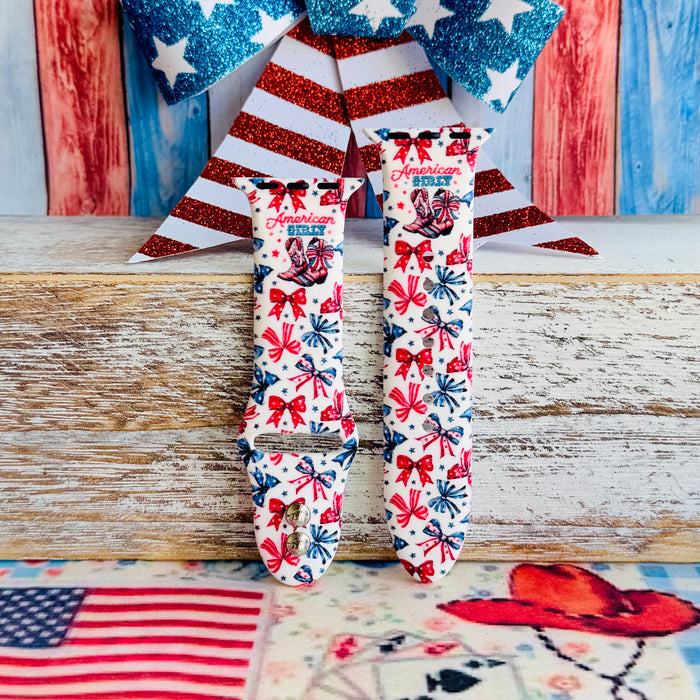 American Girly silicone Apple Watch band with red and blue coquette bows, mini cowboy boots, stars, and "American Girly" script on a white base, shown displayed and worn on Apple Watch against a patriotic 4th of July backdrop.