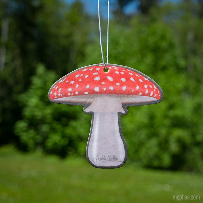 The Aromatic Mushroom Air Freshener, adorned with a red cap and white spots, is displayed outdoors against a softly blurred green background. With its gentle sandalwood fragrance, it serves as the perfect car accessory for those looking to add a natural element to their journeys.