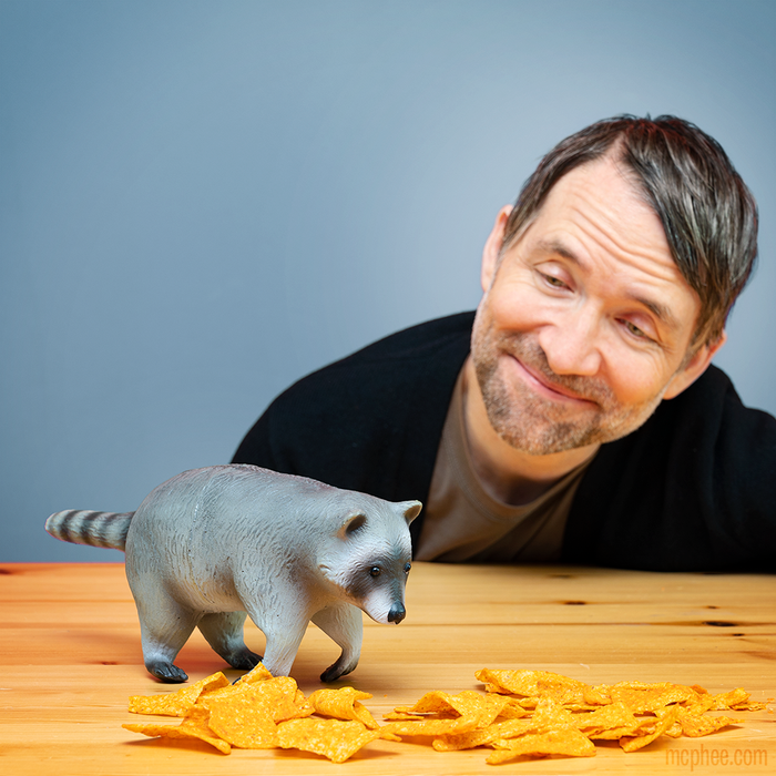 A man smiling at the Baby Raccoon Buddy, a stuffed animal with posable paws, which sits on a table surrounded by tortilla chips.
