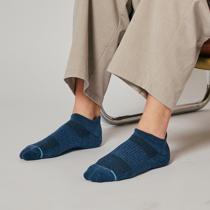 Ankle compression socks in dark blue worn with light-colored trousers, sitting on a chair.