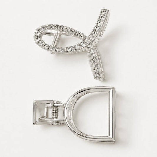 A silver-toned hair claw clip shown from two angles on a clean white background. One view shows the front with a curved open design set with clear rhinestones, and the other shows the back with a D-shaped frame and spring-loaded clasp mechanism.