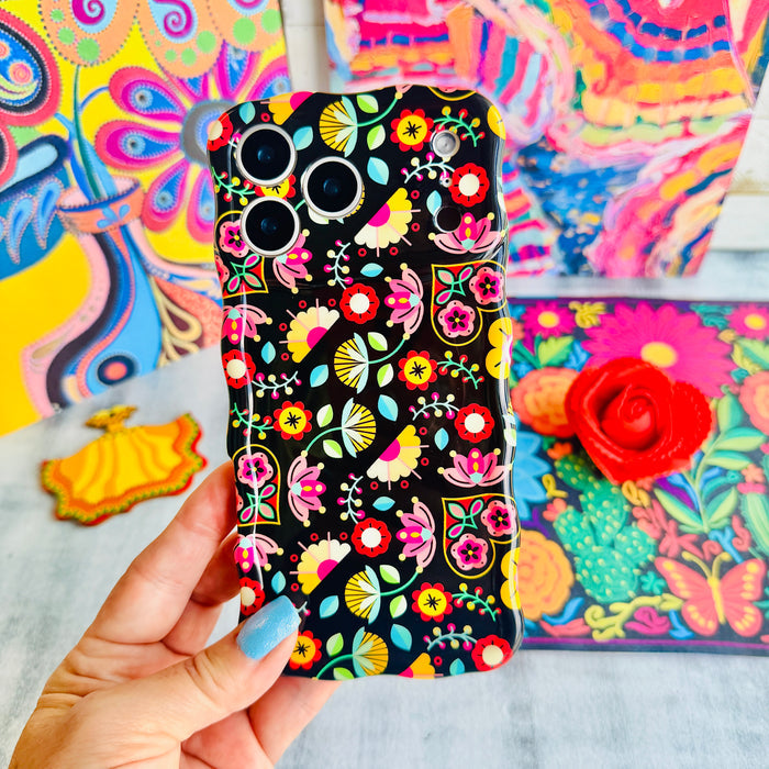 Trendy black wavy iPhone case featuring an intricate folk art botanical pattern of red flowers, magenta details, and yellow fan petals with a high-gloss finish.