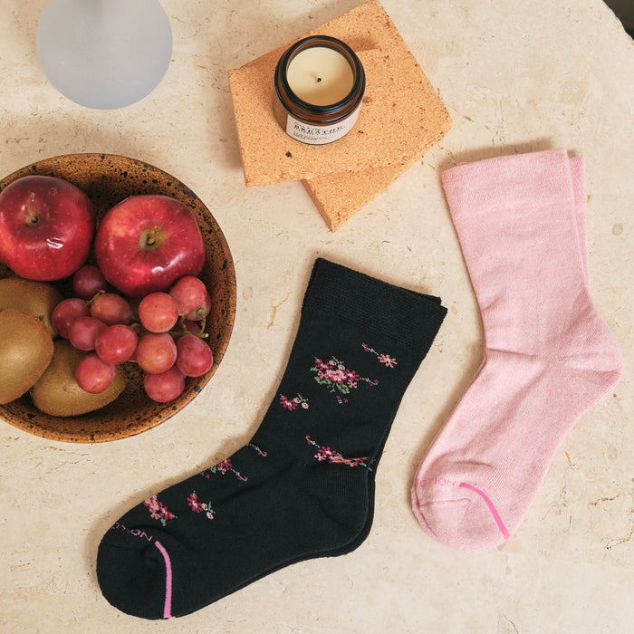 Black floral and pink half-cushion diabetic socks next to a bowl of apples and grapes, with a candle on a cork coaster.