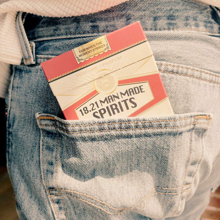 A close-up of a person wearing light-wash blue denim jeans, with a red and cream 18.21 Cologne Discovery Set