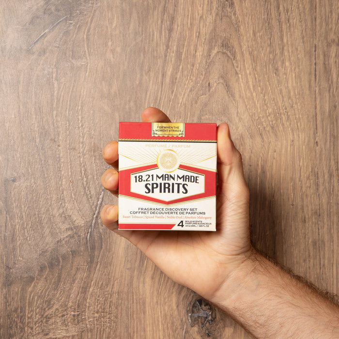 A hand holding a small box of 18.21 Minute Spirits cologne discover set  against a brown wood-grain background.