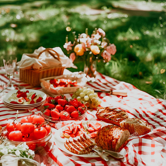 A picnic setup with a variety of food including fruits, bread, and a floral arrangement, on a checkered red and white blanket.