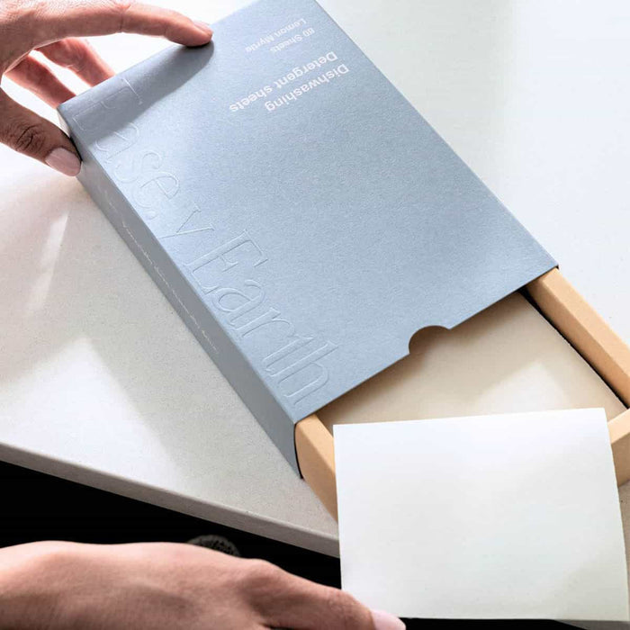 Person opening a box of dishwashing detergent sheets on a white surface