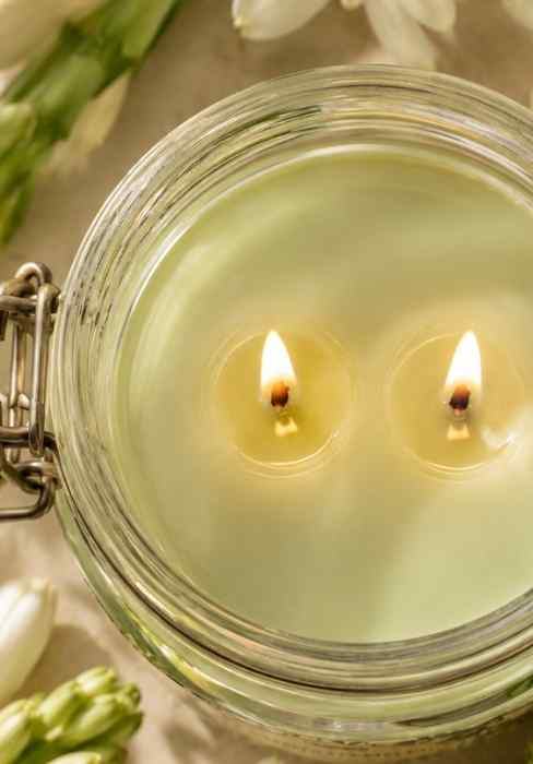 Close-up glass jar candle with lid and two lit wicks over pale green wax