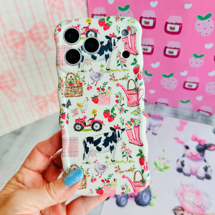 Hand holding the Farm Girl Cutie Wavy Luxe MagSafe iPhone case, showing the cream print with cows, strawberries, watering cans and a red tractor.
