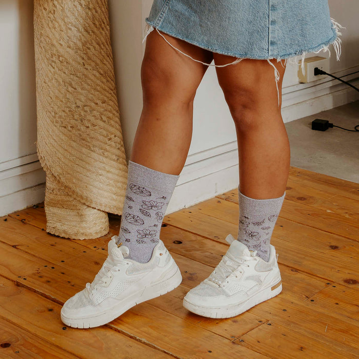 Gray socks with floral outline design worn with white sneakers and a denim skirt, standing on wooden flooring.