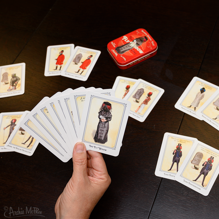 A hand holds humorously illustrated Fly Widow cards with more cards spread on the table and a red tin box nearby, reminiscent of an Old Maid game.