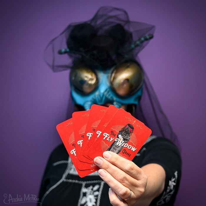 Sporting a bug mask, a person humorously displays illustrated red cards against a purple background, capturing the playful essence of the Fly Widow Game.