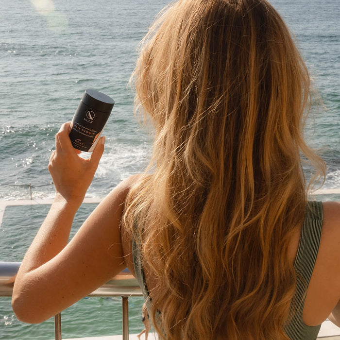 Woman with long blonde hair holding a supplement bottle by the water.