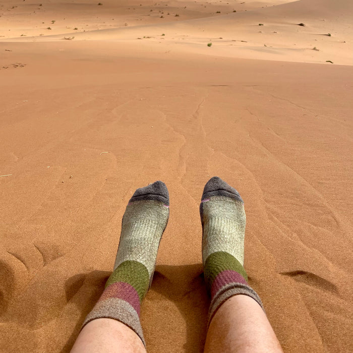 Feet wearing medium weight crew socks with a gradient design, resting on sandy terrain.