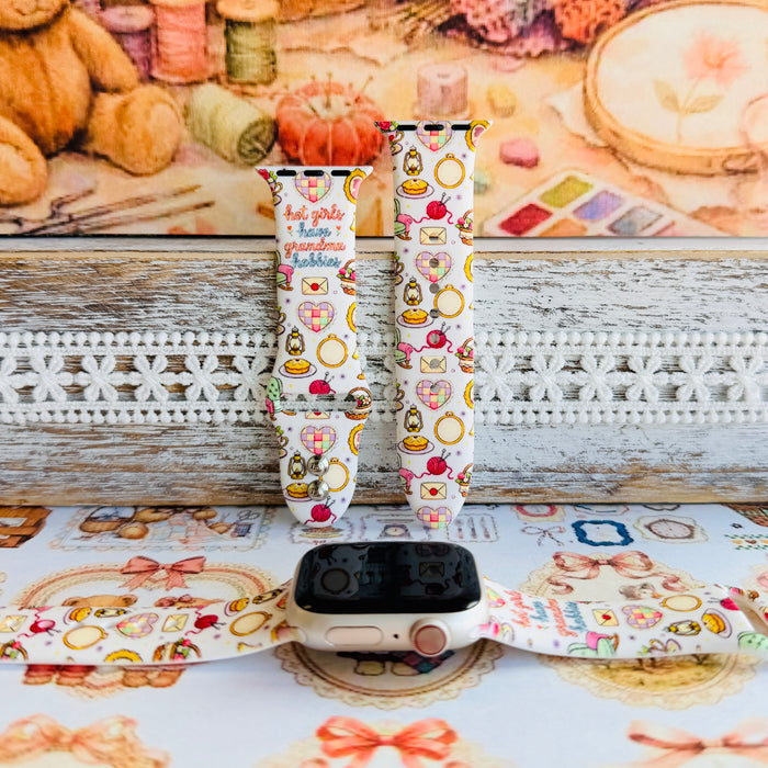 It's Giving Grandma Hobbies silicone Apple Watch band with "hot girls have grandma hobbies" script, patchwork hearts, yarn balls, knitting needles, embroidery hoops, lanterns, pies, and teacups on a cream base, shown displayed and worn on Apple Watch against a cottagecore craft backdrop.