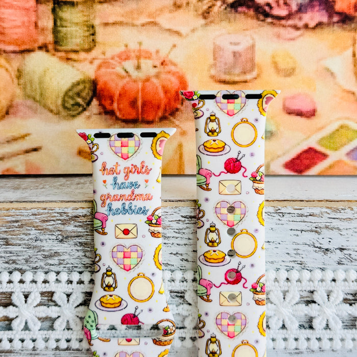It's Giving Grandma Hobbies silicone Apple Watch band with "hot girls have grandma hobbies" script, patchwork hearts, yarn balls, knitting needles, embroidery hoops, lanterns, pies, and teacups on a cream base, shown displayed and worn on Apple Watch against a cottagecore craft backdrop.