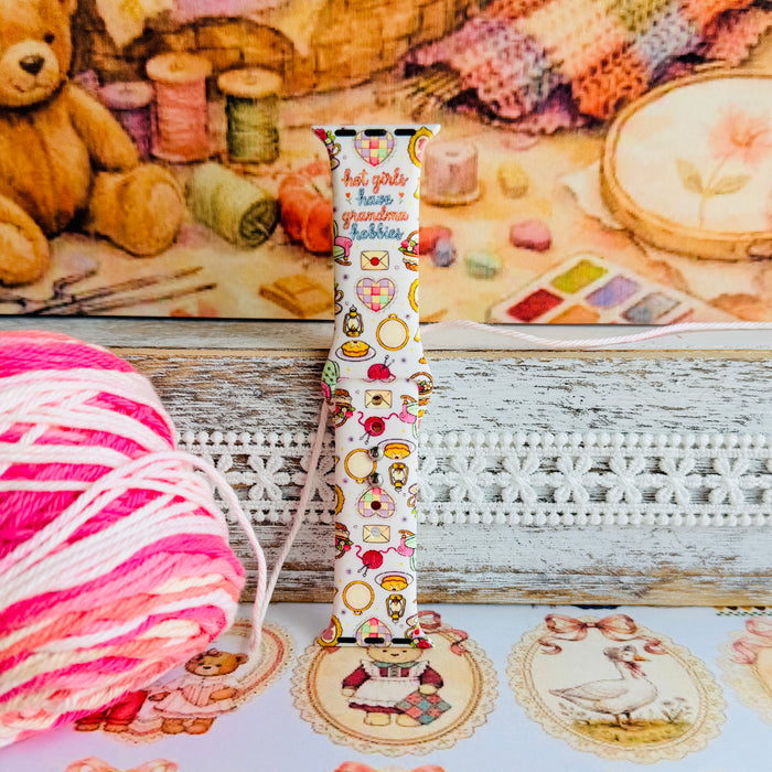 It's Giving Grandma Hobbies silicone Apple Watch band with "hot girls have grandma hobbies" script, patchwork hearts, yarn balls, knitting needles, embroidery hoops, lanterns, pies, and teacups on a cream base, shown displayed and worn on Apple Watch against a cottagecore craft backdrop.