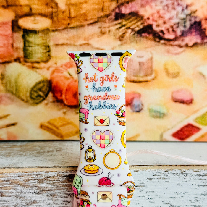 It's Giving Grandma Hobbies silicone Apple Watch band with "hot girls have grandma hobbies" script, patchwork hearts, yarn balls, knitting needles, embroidery hoops, lanterns, pies, and teacups on a cream base, shown displayed and worn on Apple Watch against a cottagecore craft backdrop.