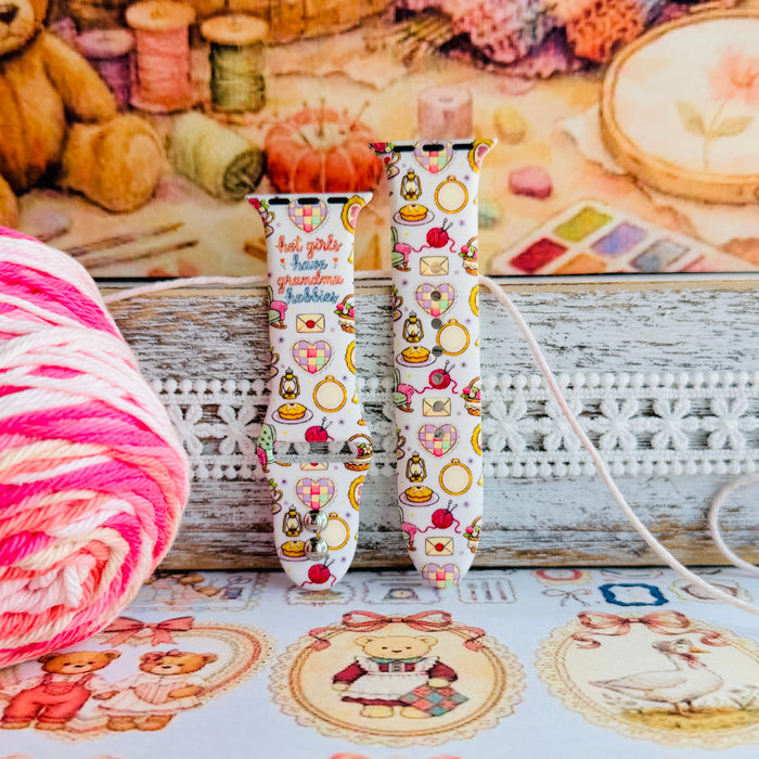 It's Giving Grandma Hobbies silicone Apple Watch band with "hot girls have grandma hobbies" script, patchwork hearts, yarn balls, knitting needles, embroidery hoops, lanterns, pies, and teacups on a cream base, shown displayed and worn on Apple Watch against a cottagecore craft backdrop.