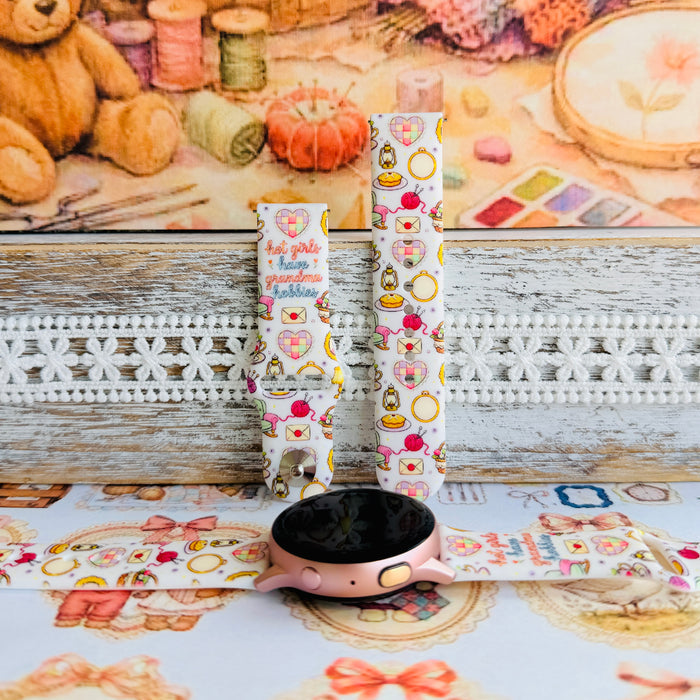 It's Giving Grandma Hobbies silicone Samsung Galaxy Watch band with "hot girls have grandma hobbies" script, patchwork hearts, yarn balls, knitting needles, embroidery hoops, lanterns, pies, and teacups on a cream base, shown displayed and worn against a cottagecore craft backdrop.