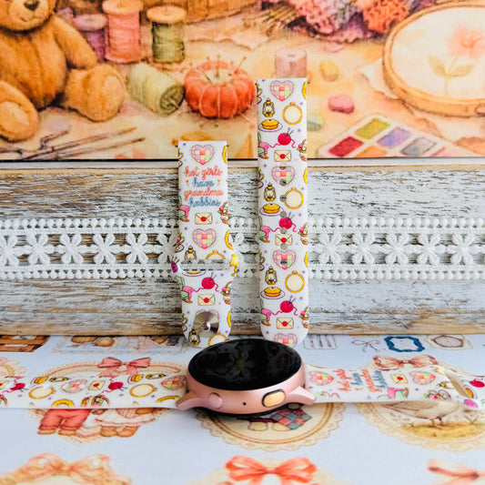 It's Giving Grandma Hobbies silicone Samsung Galaxy Watch band with "hot girls have grandma hobbies" script, patchwork hearts, yarn balls, knitting needles, embroidery hoops, lanterns, pies, and teacups on a cream base, shown displayed and worn against a cottagecore craft backdrop.