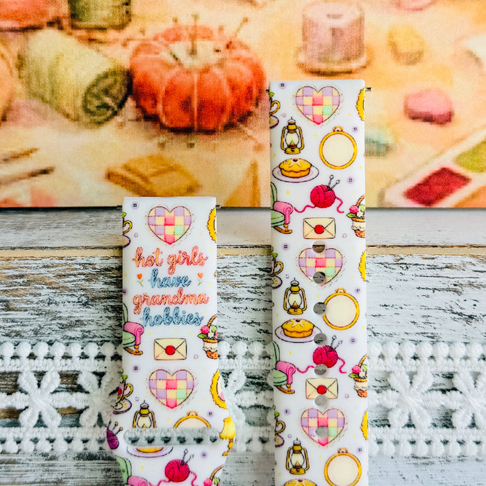 It's Giving Grandma Hobbies silicone Samsung Galaxy Watch band with "hot girls have grandma hobbies" script, patchwork hearts, yarn balls, knitting needles, embroidery hoops, lanterns, pies, and teacups on a cream base, shown displayed and worn against a cottagecore craft backdrop.