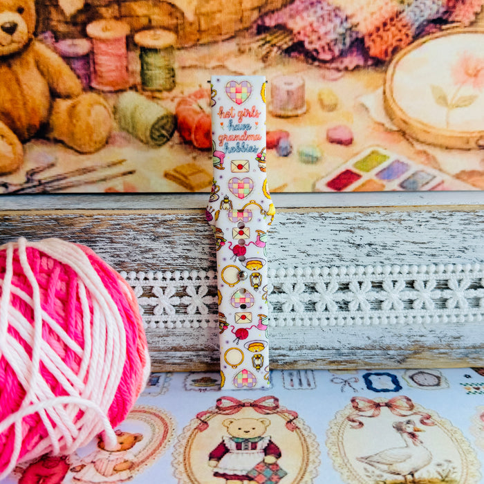 It's Giving Grandma Hobbies silicone Samsung Galaxy Watch band with "hot girls have grandma hobbies" script, patchwork hearts, yarn balls, knitting needles, embroidery hoops, lanterns, pies, and teacups on a cream base, shown displayed and worn against a cottagecore craft backdrop.