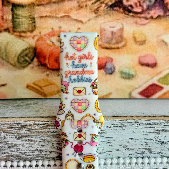 It's Giving Grandma Hobbies silicone Samsung Galaxy Watch band with "hot girls have grandma hobbies" script, patchwork hearts, yarn balls, knitting needles, embroidery hoops, lanterns, pies, and teacups on a cream base, shown displayed and worn against a cottagecore craft backdrop.