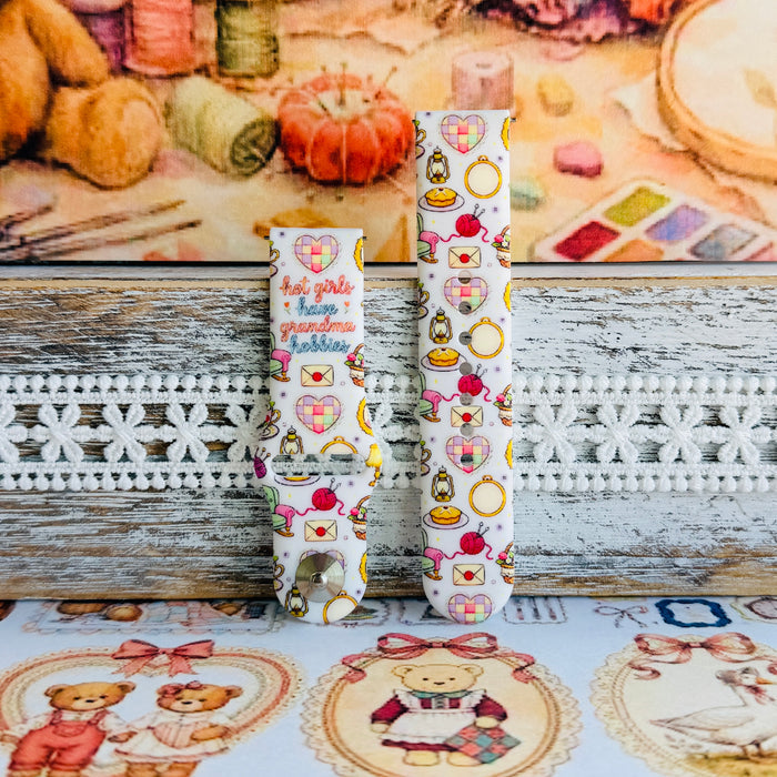 It's Giving Grandma Hobbies silicone Samsung Galaxy Watch band with "hot girls have grandma hobbies" script, patchwork hearts, yarn balls, knitting needles, embroidery hoops, lanterns, pies, and teacups on a cream base, shown displayed and worn against a cottagecore craft backdrop.