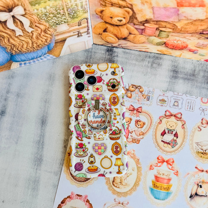 It's Giving Grandma Hobbies Wavy Luxe iPhone case with the matching MagSafe grip ring attached, styled with a teddy bear and embroidery hoop backdrop.