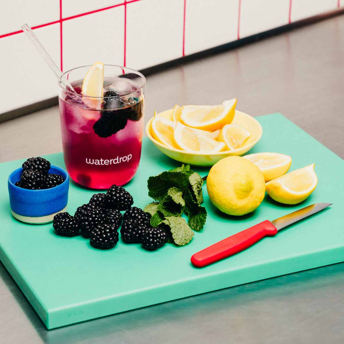 Glass of pink drink with lemon slices, blackberries, and a knife on a green cutting board.