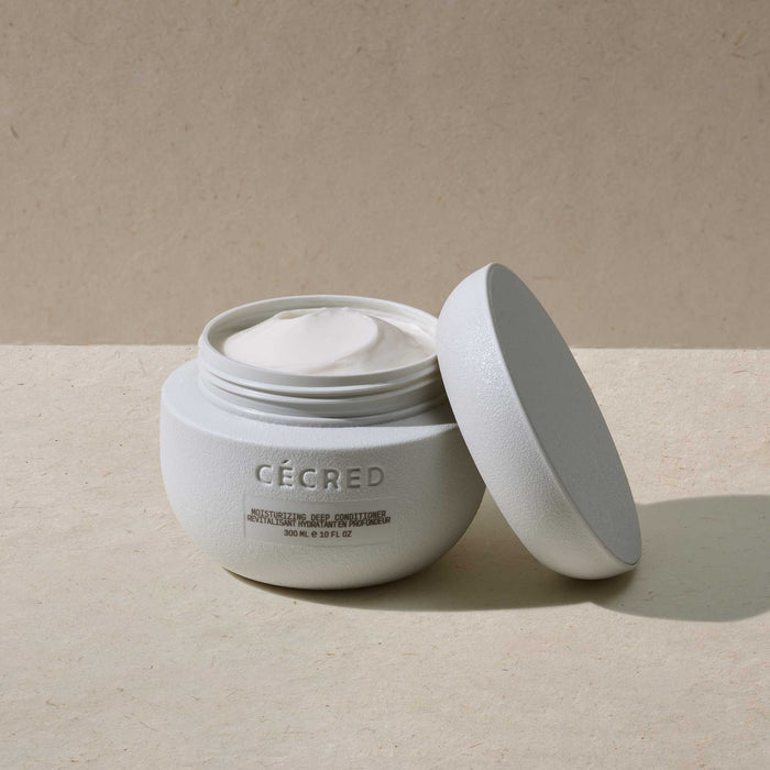 Cécred Moisturizing Deep Conditioner 300ml jar open with lid leaning against it, showing creamy white formula against a warm neutral textured surface